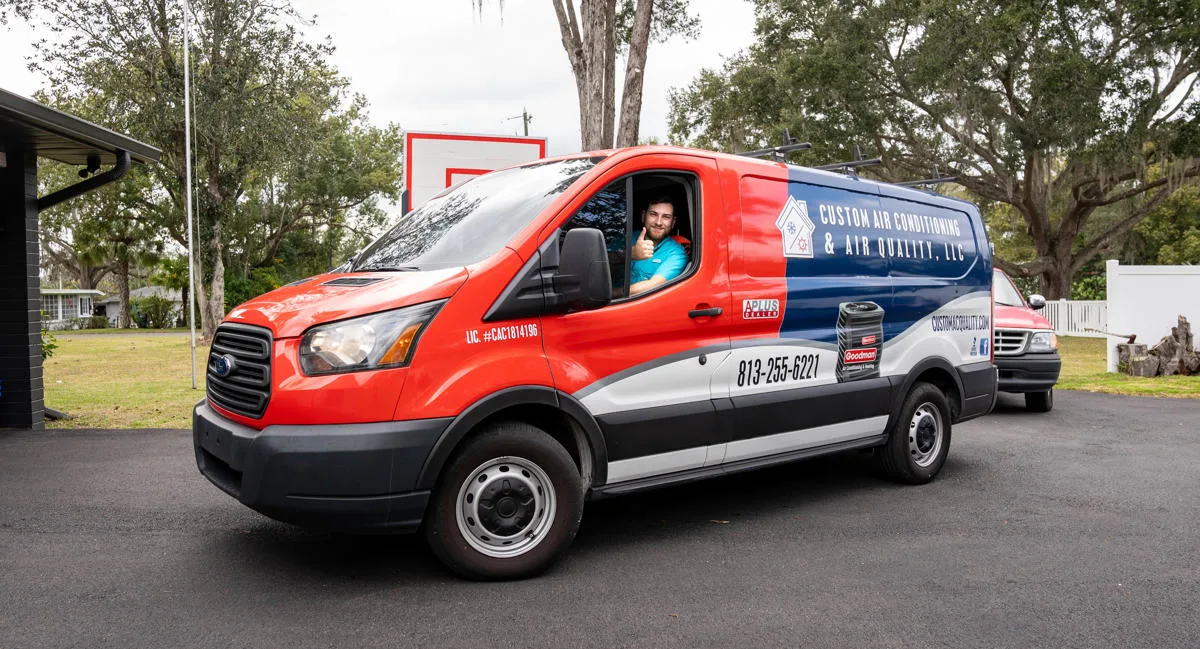 A Custom Air Conditioning Air Quality technician gives a thumbs up from the drivers seat of a branded company van showcasing the companys logo contact number and services
