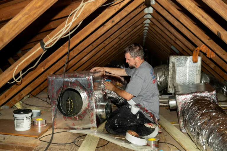 A technician works in a large attic sealing and insulating HVAC ductwork with metallic tape. The technician is kneeling next to a large insulated air handler