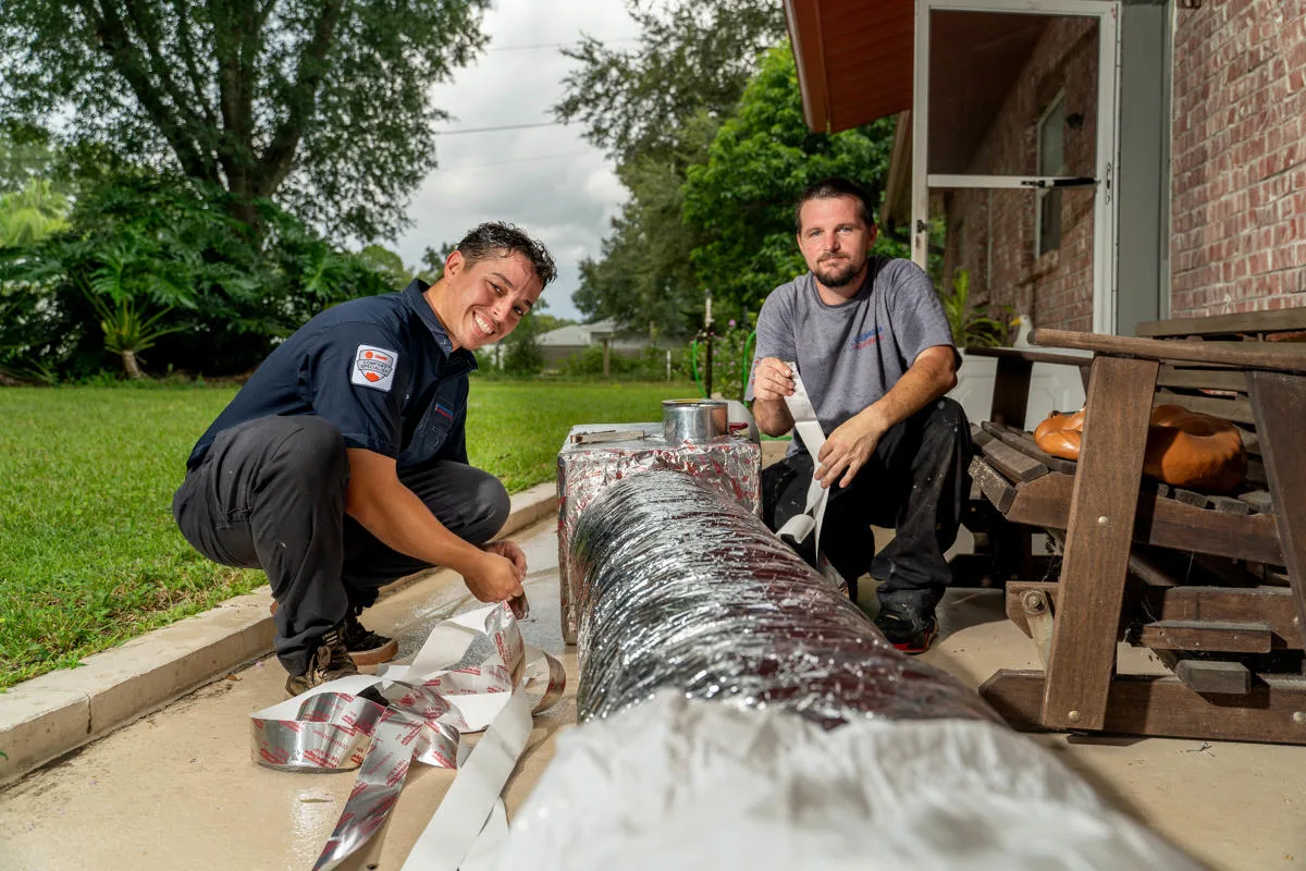 Two Custom Air Conditioning Air Quality technicians work on wrapping and sealing an insulated air duct outside a home both smiling and focused on the task