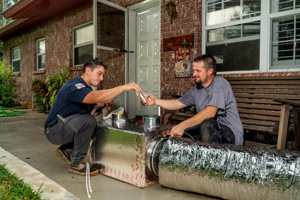 Two HVAC technicians work together outdoors on a large insulated air duct. One technician hands a pair of pliers to the other as they collaborate on assembling ductwork